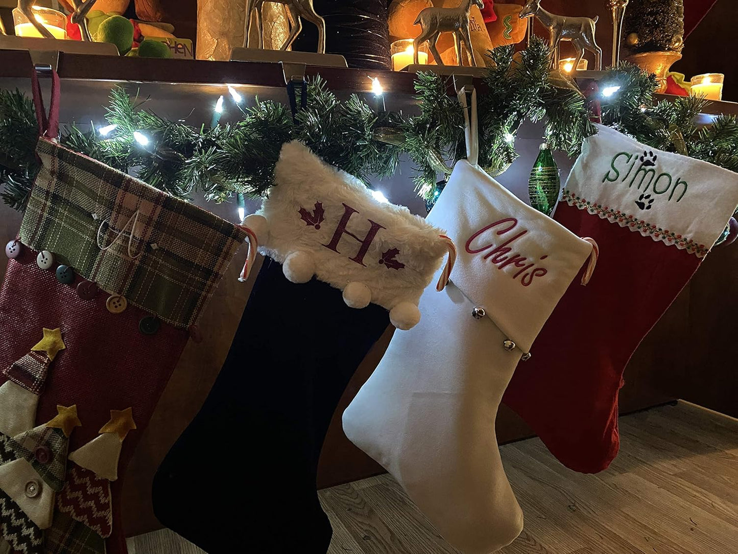 Four Personalized Christmas Stockings, including an extra large jute and red one, hang on a garland and light-decorated mantel. Three stockings read H, Chris, and Simon. Candles, ornaments, and reindeer figurines add a festive touch above.