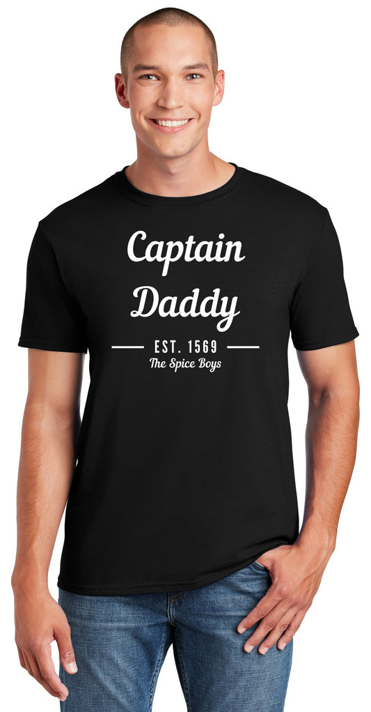 A smiling man with short hair wears the Spice Boys Captain Daddy - Gildan Softstyle T-Shirt (64000), a black, modern classic fit tee made from ring spun US cotton, featuring white "Captain Daddy EST. 1569 The Spice Boys" text. One hand in his jeans pocket.