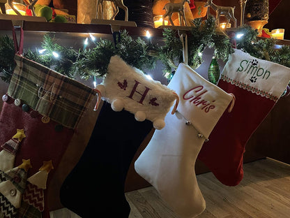 Four Personalized Christmas Stockings, including an extra large jute and red one, hang on a garland and light-decorated mantel. Three stockings read H, Chris, and Simon. Candles, ornaments, and reindeer figurines add a festive touch above.