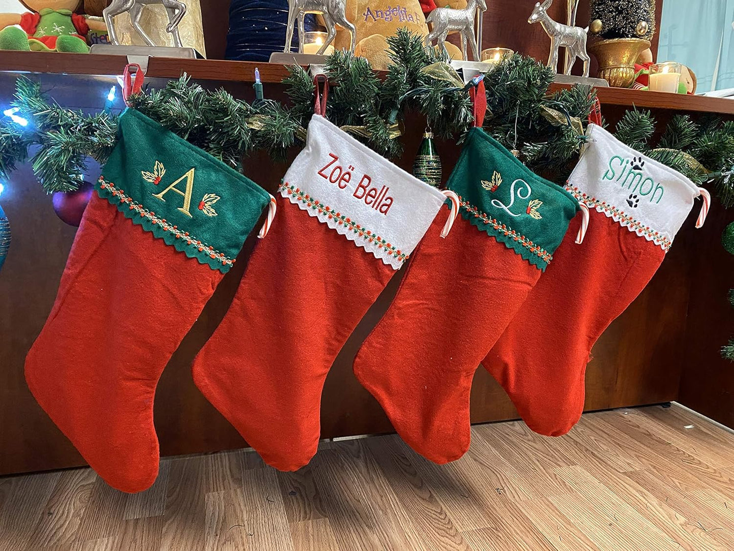 Four Personalized Christmas Stockings in red and green felt, each with a custom name—A, Zoë Bella, S, and Simon—hang from a garland-decorated mantle. Festive lights and decor enhance the holiday warmth in the background.