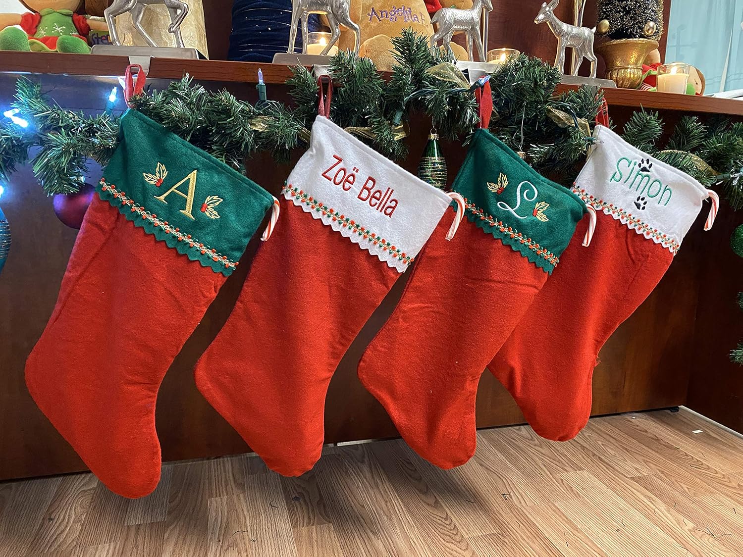 Four Personalized Christmas Stockings in red and green felt, each with a custom name—A, Zoë Bella, S, and Simon—hang from a garland-decorated mantle. Festive lights and decor enhance the holiday warmth in the background.