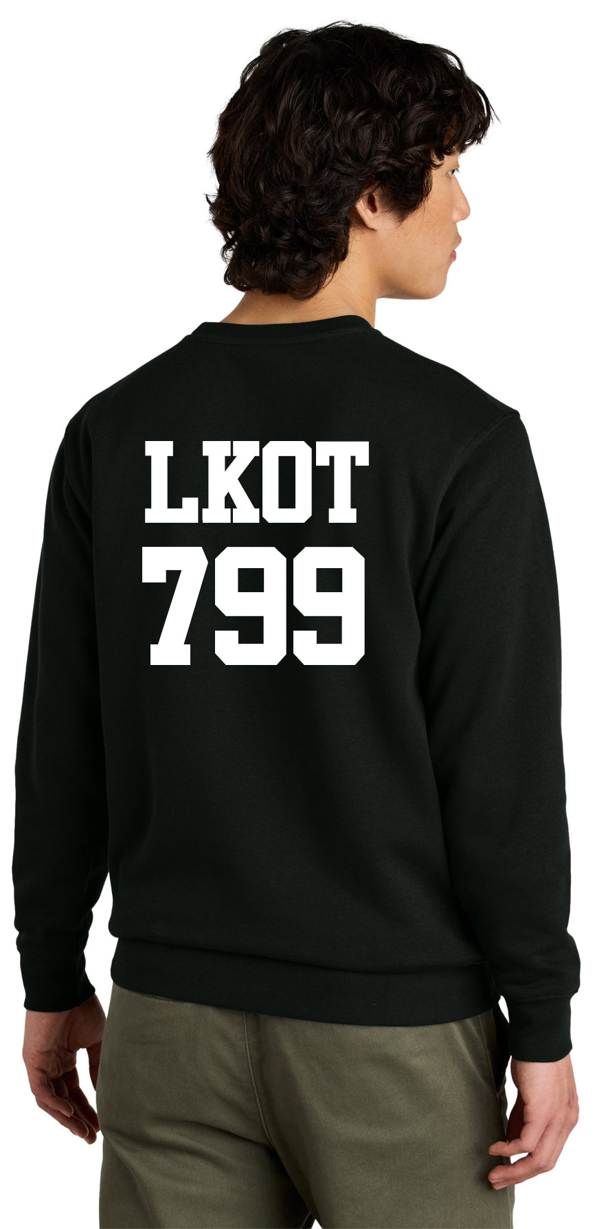 A person with short, dark hair is seen from behind wearing the LKOT Crewneck Sweatshirt — Adult in black, featuring bold white "LKOT 799" text, styled like a collegiate crewneck. They pair it with olive green pants.