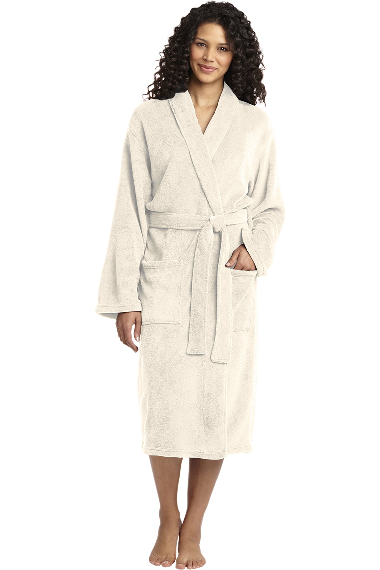 A woman with curly hair stands barefoot, smiling and facing forward in the Personalized Plush Robe—a light-colored, knee-length style with a shawl collar, belt, and two front pockets for cozy luxury.