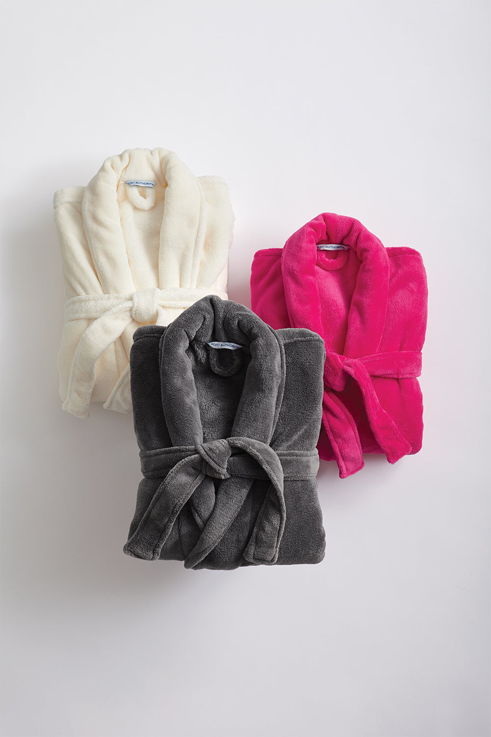 Three Personalized Plush Robes in cream, bright pink, and dark gray are displayed on a white background. Each features a plush shawl collar, matching belt tied at the front, and offers cozy luxury for your home.