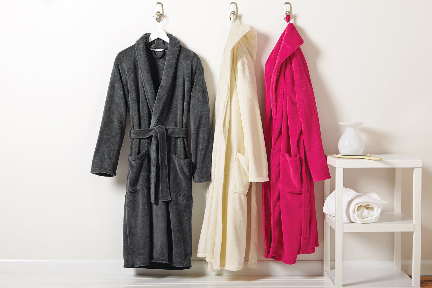 Three Personalized Plush Robes—dark gray with a shawl collar, cream, and bright pink polyester fleece—hang on hooks against a light wall. A white shelf nearby displays a folded towel and a white vase.