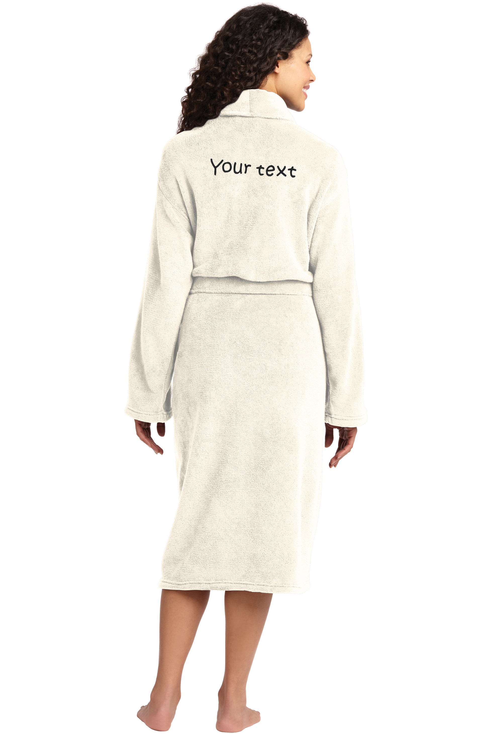 A woman with curly hair stands barefoot, wearing the Personalized Plush Robe with a plush shawl collar. "Your text" is embroidered in black on the back as she faces away from the camera.
