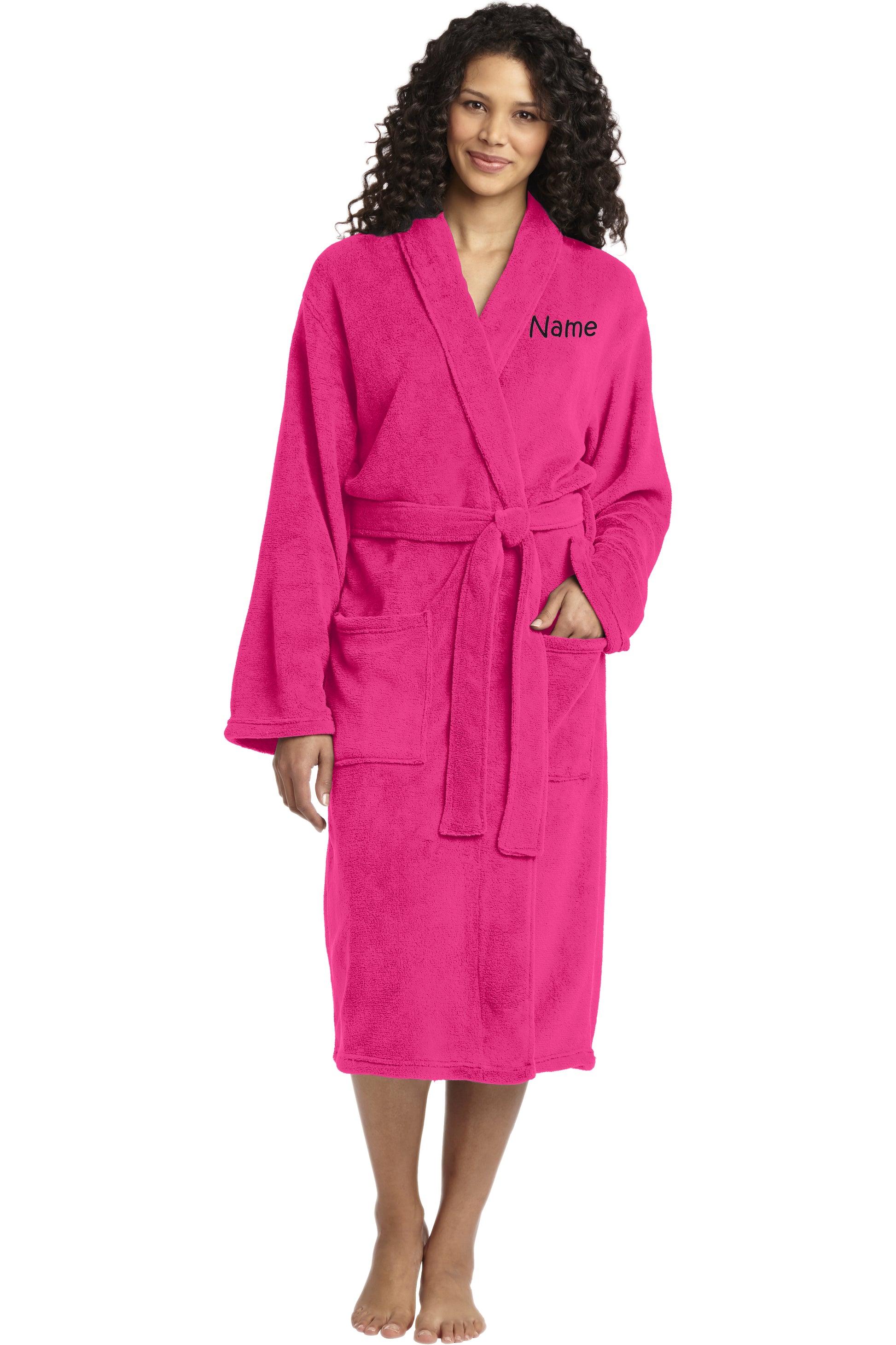 A woman with curly hair wears a bright pink Personalized Plush Robe featuring a waist tie, large front pockets, and her name embroidered on the upper left. She is barefoot and smiling at the camera.