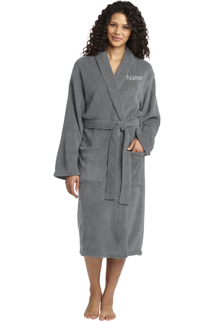 A smiling woman with curly hair wears the Personalized Plush Robe— a long, gray polyester fleece robe with a belt, large front pockets, and "Name" embroidered on the left chest. She stands barefoot.