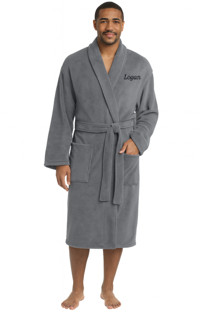 A man smiles barefoot in a Personalized Plush Robe made of cozy gray polyester fleece, featuring a shawl collar, belt, two front pockets, and “Logan” embroidered on the left chest. One hand rests casually in his pocket.